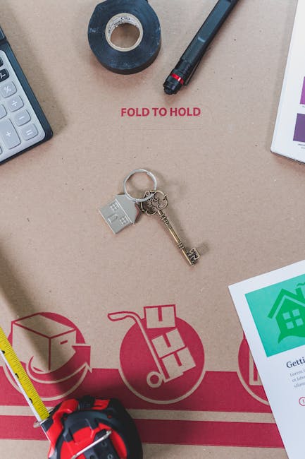 A close-up view of a beige cardboard moving box with red printed instructions and graphics, placed inside a home during a furniture transport and packing process. Positioned on the box are various items including a silver key attached to a keyring with a small house-shaped charm, a roll of black packing tape, a black marker with a red cap, and a partial view of a white electronic device or tablet on the right. To the left, a yellow measuring tape is partially visible, and a grey calculator is seen at the top left corner. The scene captures elements typical of home relocation preparations, with the box serving as a base for organizing and packing belongings, supported by the presence of moving and packing tools. Man With a Van Palmers Green's removals services are suggested by these visual cues related to house moving and furniture transport.