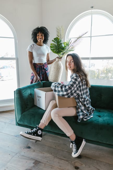 Two women are indoors in a bright room with large arched windows, engaged in a home relocation or packing process. One woman with curly hair and a casual white t-shirt is standing, holding a large dec
