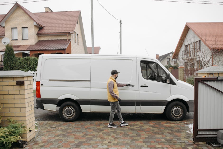 A man wearing a yellow and grey jacket, grey trousers, and a black face mask is standing on a brick-paved driveway, opening the sliding door of a white commercial van parked in front of a residential property in Palmers Green. The van is an urban cargo vehicle with a black stripe along the side, positioned close to the side of the house, which has a yellow brick exterior and a red-tiled roof. Inside the driveway, there are several cardboard boxes, plastic-wrapped items, and a piece of furniture covered with protective blankets, indicating the process of packing or home relocation. The scene is outdoors during daylight with overcast skies, and neighboring houses with similar red tiles and garden areas are visible in the background. The image clearly shows the loading process involved in furniture transport and removal services provided by Man With a Van Palmers Green, reflecting aspects of packing, moving logistics, and vehicle loading for house removals.