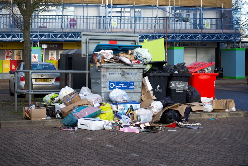 An overflowing set of large waste bins and recycling containers is positioned on the pavement outside a commercial building in a residential area, with various types of waste such as paper, cardboard boxes, plastic bags, and packaging materials scattered on the ground nearby. The waste bins include one grey mixed paper and card container, a black bin, and a bright red recycling bin, some of which are open and filled with assorted refuse. The surrounding environment features parked cars, a metal railing, and a building with signage and blue scaffolding indicating ongoing construction or maintenance work. The scene is illuminated by daylight, with an overall impression of clutter and excess rubbish, typical of waste disposal situations related to home relocation or moving services carried out by Man With a Van Palmers Green, supporting efficient furniture transport and packing logistics for local removals.