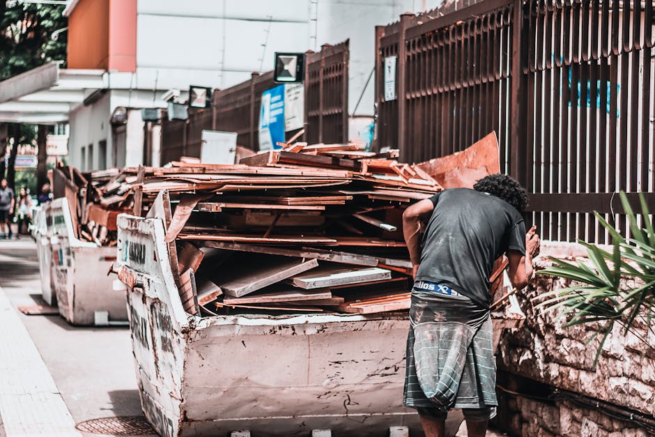 A man with curly dark hair, wearing a black T-shirt and grey shorts with a plaid pattern, stands next to a large, open skip filled with construction waste and bulky debris, including wooden planks, tiles, and various building materials. The skip is positioned on a pavement alongside a brown metal fence, with a residential or commercial building in the background, partly visible with orange walls and white window frames. Several large white containers are visible behind the skip. The scene appears to be part of a home relocation or clearance process, involving loading or disposal of bulky waste materials, with natural daylight illuminating the area. This image exemplifies typical logistics involved in house removals and furniture transport, where waste removal is a key step facilitated by specialist services such as Man With a Van Palmers Green.
