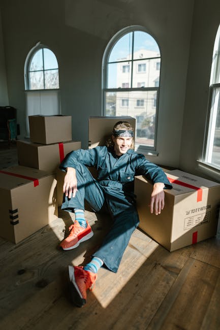 A young man wearing a dark blue work uniform, a headband, and bright orange sneakers is sitting on a wooden floor inside a room with large arched windows that let in natural light. He is surrounded by several cardboard boxes sealed with red and black packing tape, some stacked on top of each other, ready for a home relocation. The room appears empty aside from the boxes, with minimal furnishings. Outside the windows, a building and trees are visible. This scene captures the loading and packing phase of a house removal, illustrating furniture transport and moving logistics. The presence of boxes and the indoor setting emphasize the packing process involved in professional removals services offered by Man With a Van Palmers Green, highlighting their expertise in quick and efficient moving solutions.