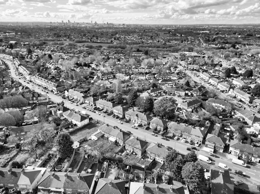 Black and white aerial photograph displaying a residential neighbourhood with rows of terraced houses, detached, and semi-detached homes, along with tree-lined streets and parked cars. In the foreground, various backyards with visible fences, sheds, and garden furniture are seen, indicating a typical suburban area. The middle ground features houses with pitched roofs, sometimes with driveways and small front gardens. A main road runs through the scene, with several cars parked along the curbs and moving vehicles on the street. Beyond the residential area, open fields and further housing developments extend towards the horizon, under a sky filled with scattered clouds. The image captures the extensive scope of the neighbourhood, suggesting a well-established community suitable for home relocations involving furniture transport and moving logistics, typical of house removals by companies like Man With a Van Palmers Green.