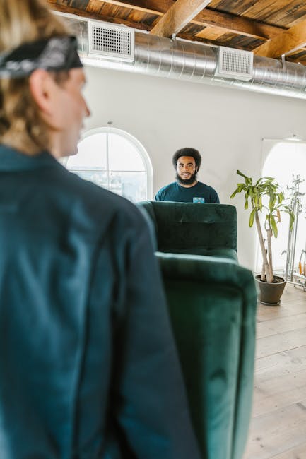 A man wearing a dark jacket and headband is inside a room, partially visible, lifting or positioning a large, dark green upholstered armchair during a home relocation process. The room features a wooden ceiling with exposed beams and metallic ductwork, with natural light coming through a rounded window. In the background, another man with curly hair and a beard is standing near a potted plant, possibly overseeing the moving activity. The setting appears to be part of an indoor furniture transport or packing and moving operation, with the environment indicating careful handling of household items. The scene is part of a professional removal service, such as Man With a Van Palmers Green, involved in furniture transport and house removals, supporting the logistics of a home move against a minimalist interior backdrop.