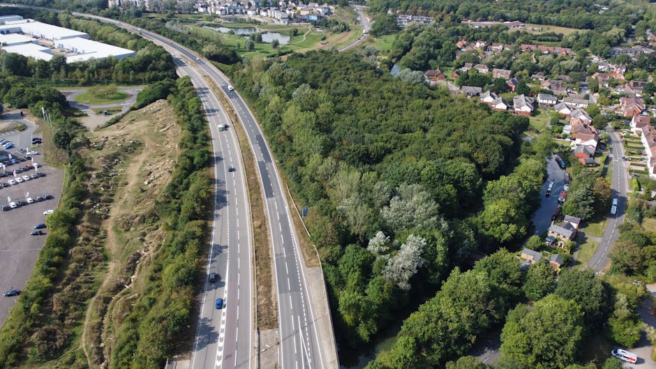 An aerial view showing a dual carriageway with multiple lanes in each direction, surrounded by lush green trees and dense woodland on both sides. To the left of the road, there is an open parking area filled with numerous parked vehicles. On the right side, residential houses with gardens are visible, arranged along curved streets that follow the natural landscape. The scene is taken during the daytime with bright, natural lighting, highlighting the contrast between the urban infrastructure and the surrounding greenery. This image illustrates the typical environment associated with house relocations and moving logistics in suburban areas, emphasizing the importance of planning when transporting furniture and boxes through such landscapes, as supported by services like Man With a Van Palmers Green.