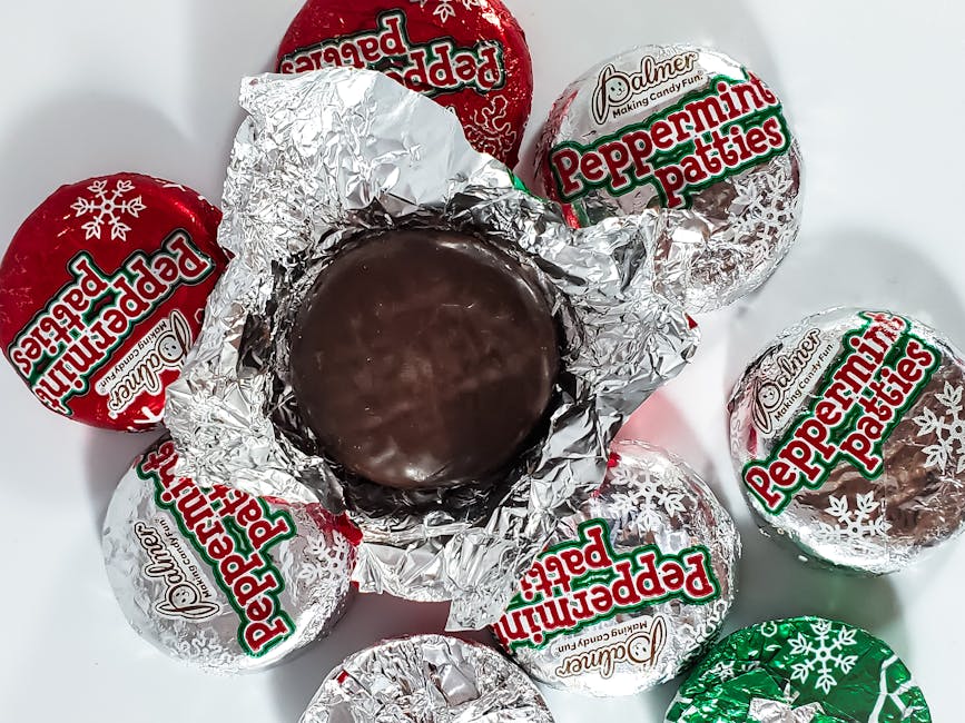 Photograph showing multiple Peppermint Patties wrapped in clear and coloured foil wrappers, with one unwrapped peppermint candy in the centre. The candies are arranged on a white surface, likely for packing or transport, with some wrappers displaying the 'Palmer' brand logo. The unwrapped candy has a smooth, round chocolate coating, suggesting it is part of a packing and moving process involving confectionery or snack items, possibly during home relocation. Visible items include the foil wrappers, with some crinkled from handling, and the unwrapped peppermint candy placed atop a piece of crumpled foil. The scene reflects careful handling of confectionery packaging, typical during packing procedures conducted by removals specialists such as Man With a Van Palmers Green, who manage various household items, including snacks, during furniture transport or packing and moving tasks.