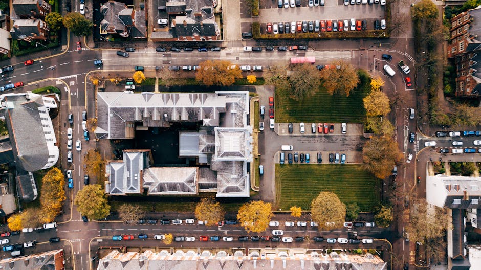 An aerial view of a city block showing a large multi-storey building with a grey, pitched roof and several smaller attached sections. Surrounding the building are trees with autumn foliage, parking areas filled with numerous cars, and narrow streets with parked vehicles. Adjacent to the property are paved pathways and green lawns, with some staff members or movers not visibly present in the image. The scene captures the environment where house removals and furniture transport operations may take place, illustrating the parking and access conditions relevant to home relocation services by Man With a Van Palmers Green.