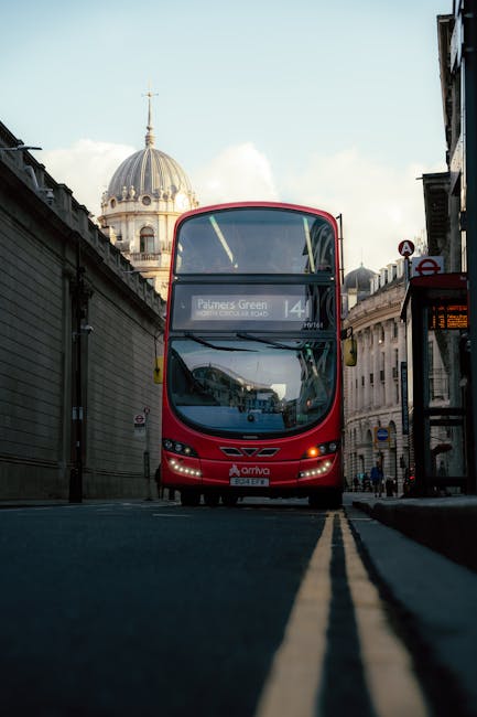 A red double-decker bus operated by Arriva is positioned in the middle of a narrow street in central Palmers Green, with its front facing the camera. The bus displays route number 141 and the destination 'Palmers Green' on the electronic sign above the windshield. To the left and right, there are tall, grey stone buildings with traditional architecture, some with decorative details and windows, framing the scene. In the background, a historic domed building with a golden weather vane on top is visible, set against a partly cloudy sky. The street surface is paved with dark asphalt, featuring bright yellow double lines running along the centre. To the right, a bus stop shelter with a glass panel and timetable display can be seen, and a small group of pedestrians are visible further down the street. During home relocation or furniture transport, this urban setting illustrates the typical environment where Man With a Van Palmers Green may operate, involving loading and unloading processes in city streets.