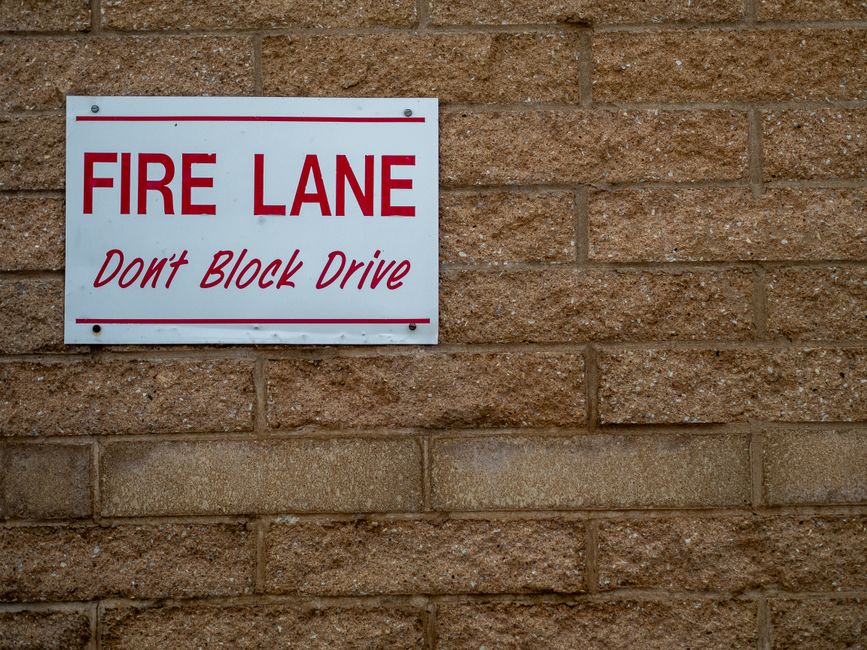 A white rectangular sign mounted on a brown brick wall, displaying the text 'FIRE LANE' in bold red letters, with a smaller message underneath in red cursive that reads 'Don't Block Drive'. The sign is positioned at eye level and serves as a reminder for parking restrictions in areas designated for emergency access. The brick wall behind the sign has a rough texture with visible mortar joints and some variation in color. This setting is relevant for house removals and home relocation services, such as those offered by Man With a Van Palmers Green, especially when navigating street-closure rules for efficient furniture transport and loading processes during local moving operations.