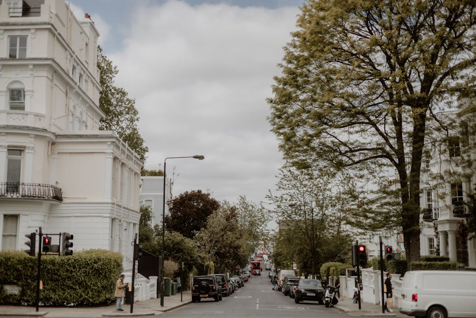 A wide view of a residential street showing white terraced Victorian-style houses with decorative facades and balconies on both sides. Several parked cars and vans line the curb, including a white van prominently visible on the right side, which appears to be part of a home relocation or furniture transport process. The street is populated with a few pedestrians, some standing near the pavement and others walking, possibly involved in moving activities. Tall trees with green foliage shade the scene, and the sky is overcast with grey clouds. Streetlights and traffic signals are visible at the intersection, with a red light illuminated, indicating stationary traffic. The overall scene suggests an active moving or packing and moving operation taking place, with the presence of moving equipment and vehicles associated with professional removals services, like those provided by Man With a Van Palmers Green, supporting home relocation tasks efficiently.